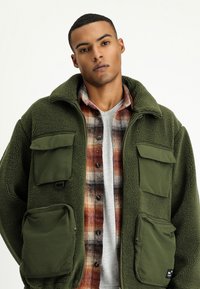 Zielona polarowa kurtka z zamkiem błyskawicznym i zatrzaskami, z wieloma przednimi kieszeniami i wysokim kołnierzem, nakładająca się na koszulę w kratę.