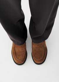 Καφέ μοκασίνια από σουέντ με στρογγυλεμένη μύτη, διακοσμητικό ράψιμο και μαύρη καουτσούκ σόλα, συνδυασμένα με σκουρόχρωμο, υφασμάτινο παντελόνι.