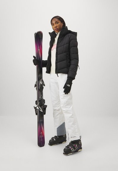 Εξοπλισμός σκι που περιλαμβάνει μια μαύρη γεμάτη μπουφάν, λευκά αδιάβροχα παντελόνια, μωβ σκι και μαύρα μποτάκια σκι, συμπληρωμένα με μαύρα γάντια.