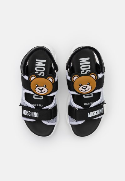 Sandálias em malha preta e branca com um acessório em forma de urso, fechos de Velcro e um logótipo na sola, apresentando uma sola espessa de borracha branca.