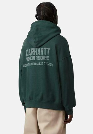 Persona con cabello trenzado vistiendo una sudadera con capucha verde oscuro con el texto "Carhartt Work in Progress Detroit Michigan" en la espalda y pantalones claros.