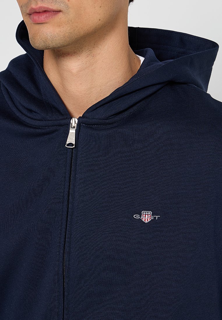 Mörkblå huvtröja med silverfärgad dragkedja, mjukt material och broderad logotyp på bröstet. Enkelt mönster utan ytterligare mönster.