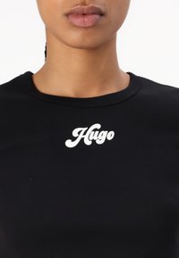 Juodas medvilninis marškinėlis su gumine tekstūra, ant krūtinės srities esantis baltojo logotipo "Hugo" stilizuotu šriftu.