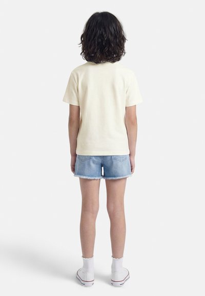 Παιδί με σκούρα, σγουρά μαλλιά μέχρι τους ώμους στέκεται με την πλάτη προς τα πίσω, φοράει κρεμ κοντομάνικο πουκάμισο, τζιν σορτς, λευκές κάλτσες και αθλητικά παπούτσια.