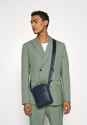 Borsa a tracolla in pelle blu navy con tasca frontale e tracolla regolabile, indossata su un completo verde chiaro. Texture liscia, forma rettangolare.