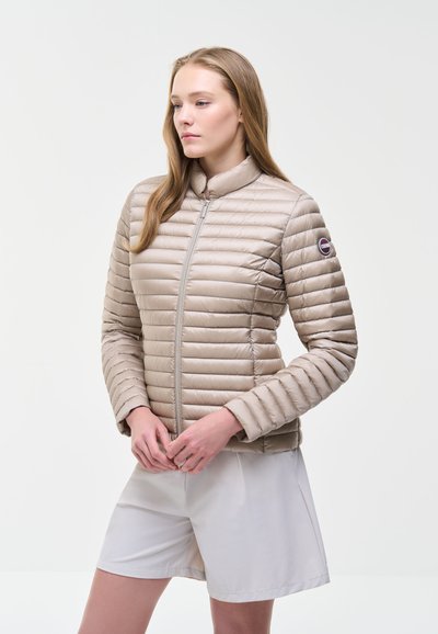 Femme aux longs cheveux châtain clair portant une veste matelassée beige et un short blanc, debout devant un fond clair uni.