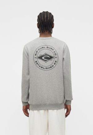Personne aux cheveux tressés portant un sweat-shirt gris avec un logo circulaire "Rip Curl Surf Co Established 69" dans le dos, associé à un pantalon blanc.