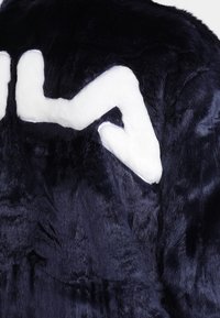 Marinblå fuskpälsjacka med en mjuk vit logotyp i form av en zigzag på ryggen. Mjuk textur med en slät yta.