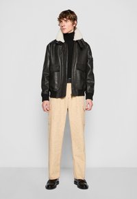 Black leather bomber jacket with a white shearling collar, zip front, two front pockets; paired with beige patterned cargo pants and black shoes.