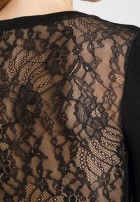 Černý krajkový cardigan s květinovými vzory, průsvitnou texturou a pevným černým lemováním podél okrajů a rukávů.