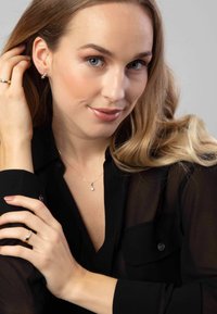Collier en métal doré avec un petit pendentif en forme d'étoile, porté sur une chemise noire transparente à boutons. Le mannequin a de longs cheveux blonds ondulés et une peau claire.