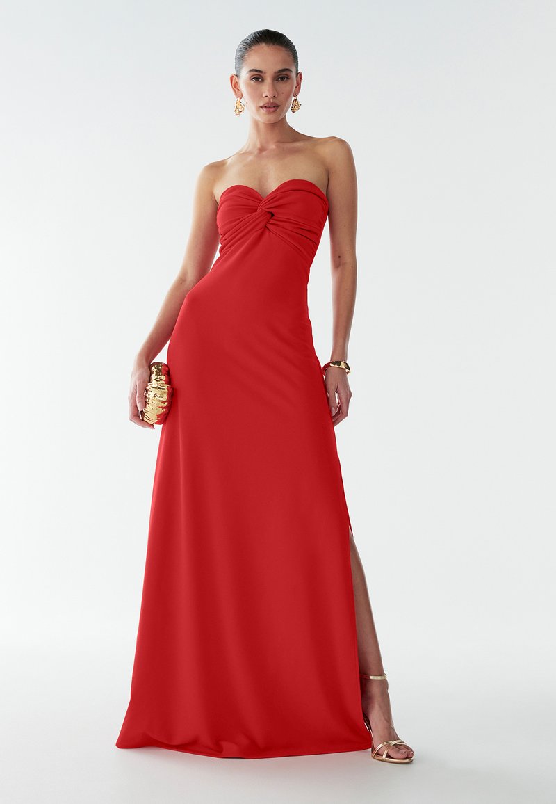 Rode strapless avondjurk met een gedraaide bustier, vloerlang ontwerp en een hoge split; gladde stof met een lichte glans; gecombineerd met een gouden clutch.