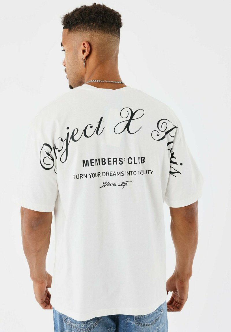 Hombre con cabello corto y rizado que lleva una camiseta blanca con texto negro "Project X Paris", "Members' Club" y frases motivacionales en la parte de atrás.
