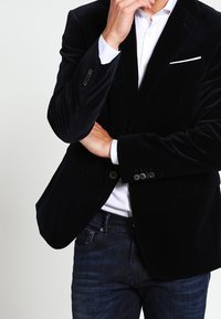 Svart sammet blazer med snedskuren slag, enkel vit fickduk och silverknapp detaljer, matchat med mörka denimjeans och en vit skjorta.