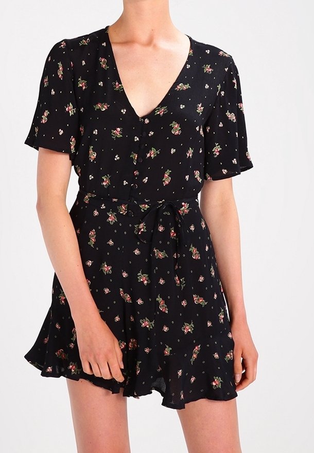 Femme portant une robe noire à manches courtes avec un motif floral, des boutons sur le devant et une ceinture nouée à la taille.