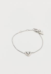 Ασημένιο βραχιόλι με μενταγιόν σε σχήμα καρδιάς, που διαθέτει ένα διαυγές κρύσταλλο στο κέντρο του, προσαρτημένο με λεπτή αλυσίδα και μικρή ετικέτα λογότυπου.