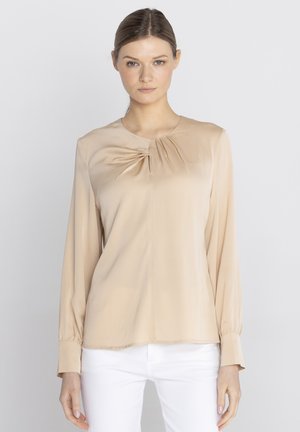 Kobieta o jasnobrązowych włosach, ubrana w beżową bluzkę z długim rękawem z fałdami przy dekolcie oraz białe spodnie, stojąca na jednolitym tle.