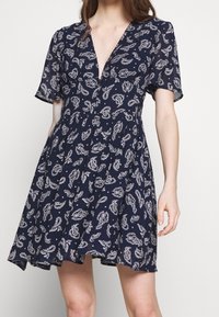 Žena v námořnickém modrém šatech s krátkým rukávem a bílým paisley potiskem, se hlubokým výstřihem do V, stojí na pozadí jednoduché světlé barvy.
