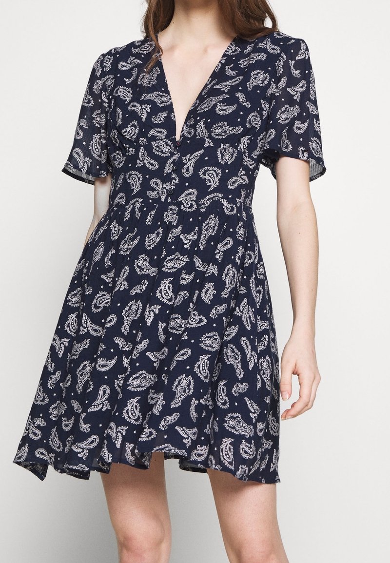 Žena v námořnickém modrém šatech s krátkým rukávem a bílým paisley potiskem, se hlubokým výstřihem do V, stojí na pozadí jednoduché světlé barvy.