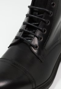 Černá kožená bota s hladkým povrchem, kulatou špičkou, šesti očky a černými tkaničkami. Obsahuje dekorativní prošívání podél špičky.