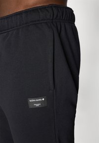 Calças de treino pretas com cintura elástica, bolsos laterais e um rótulo com a inscrição "Björk Borg, Estocolmo 1956." Textura de tecido suave.