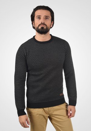 Crni teksturirani pulover s okruglim izrezom i rebrastim manžetama, uparen s bež hlačama. Uključuje malu smeđu oznaku na rubu.