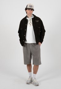 Junger Mann, der eine schwarze Cordjacke, ein weißes T-Shirt, graue kniehohe Shorts, weiße Socken, Turnschuhe, einen beigen Schal und eine Baseballkappe trägt und steht.