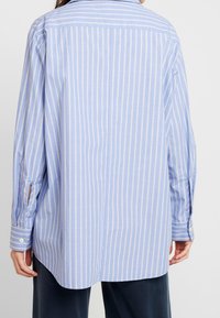 Světle modrá košile s dlouhým rukávem a bílými vertikálními pruhy, s límečkem a manžetami na knoflíky, vyrobená z hladké tkaniny.