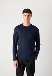 Young man wearing a navy blue long-sleeve crew neck shirt and dark pants, standing against a plain light background.