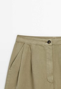 Khaki-housut, joissa on poimutettu muotoilu, nappikiinnitys ja sivutaskut. Valmistettu pehmeästä, tekstuurista kankaasta.