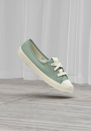 Zapatilla de lona verde claro con cordones blancos y suela de goma flotando sobre un suelo de madera gris y un fondo de cortina blanca.