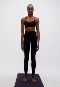 Γυναίκα με μαύρα αθλητικά ρούχα που στέκεται ξυπόλυτη πάνω σε μαύρο στρώμα γιόγκα σε απλό λευκό φόντο.