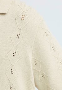 Tissu tricoté couleur crème avec un motif en losange comportant de petites ouvertures sur l'épaule et la manche d'un vêtement.