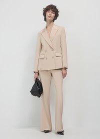 Beige doppelreihiger Blazer mit Reverskragen, kombiniert mit passenden ausgestellten Hosen. Das Modell hält eine schwarze Tasche und trägt spitze Schuhwerk.