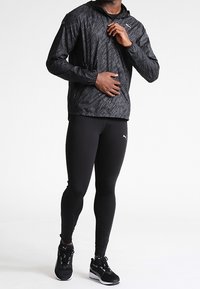 Chaqueta ligera negra con capucha y un patrón sutil, combinada con leggings ajustados negros y zapatillas deportivas. Presenta detalles con el logo de PUMA.