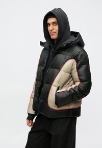 Juoda ir smėlinė pūkinė striukė su raudonu apvadu, užtrauktuku, kapišonu ir šoniniais kišenėmis. Pagaminta iš blizgaus, lengvo audinio.