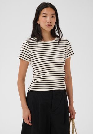 Femme aux longs cheveux noirs portant une chemise à manches courtes rayée noir et blanc et un pantalon noir, tenant la lanière d'un sac à main tissé.