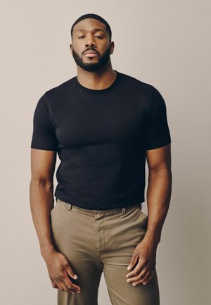 Mann mit kurzem Bart und Ohrringen, der ein schwarzes T-Shirt und beige Hose trägt und vor einem beigen Hintergrund steht.