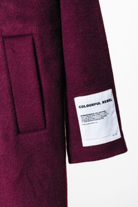 Casaco de lã burgundy com um bolso lateral e uma etiqueta branca com o nome da marca e instruções de cuidado cosidas na manga.
