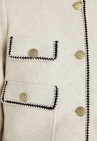 Wollen blend jas in licht-beige met zwart en wit accent. Heeft twee frontzakken met geweven gouden knopen en contrasterende stiksels.