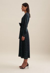 Langärmliges, dunkel-türkisfarbenes Kleid mit Leopardenmuster, mit Gürteltaille und fließendem Rock. Kombiniert mit schwarzen, geflochtenen High-Heels.