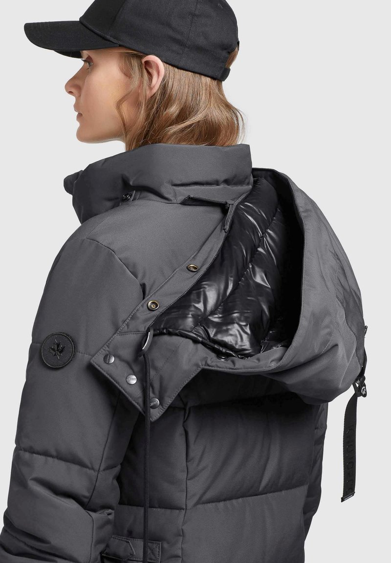 Frau in einer dunkelgrauen gepolsterten Winterjacke mit großer Kapuze und schwarzem Hut, von der Seite und von hinten vor einem hellen Hintergrund gezeigt.