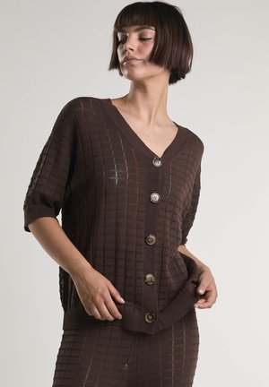 Nainen pukeutuneena ruskeaan, teksturoituun neulottuun napitettuun neuletakkiin, jossa on lyhyet hihat, sekä vastaavaan hameeseen. Hän poseeraa käsillään hameen helmassa.