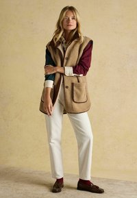 Beige Fleeceweste mit zwei Taschen, braunem Saum, getragen über einem langen grün-bordeauxfarbenen Hemd, kombiniert mit weißen Hosen und braunen Loafers.