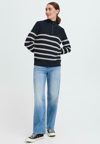 Navygestreifter Pullover mit hohem Kragen und Reißverschluss, aus strukturiertem Strickmaterial. Kombiniert mit hellblauen Schlagjeans und schwarzen Turnschuhen.