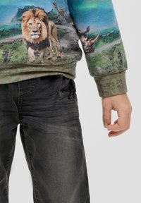 Enfant portant un pantalon gris foncé et un sweat-shirt vert avec un lion et un rhinocéros imprimés dans un design de paysage naturel.