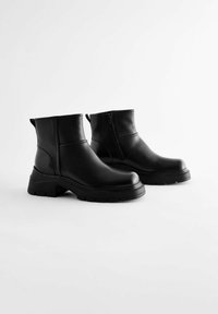 Bottines noires en cuir lisse, avec une semelle épaisse légèrement surélevée, une fermeture éclair sur le côté et un design minimaliste.