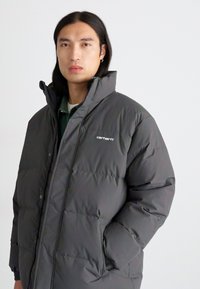 Chaqueta acolchada en gris oscuro, fabricada en nylon acolchado con un cuello alto y botones de presión. Cuenta con bolsillos laterales y un peque�ño detalle del logo.
