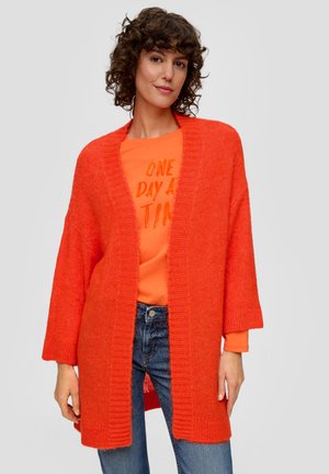Cardigan tricoté rouge-orange avec une finition texturée, à enfiler sur le devant et aux manches larges, superposé à un t-shirt graphique orange assorti.