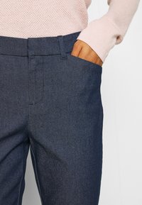 Pantalones azul marino de tejido texturizado. Cuentan con un diseño de frente plano, presillas para el cinturón y un bolsillo lateral visible con una mano descansando en su interior.